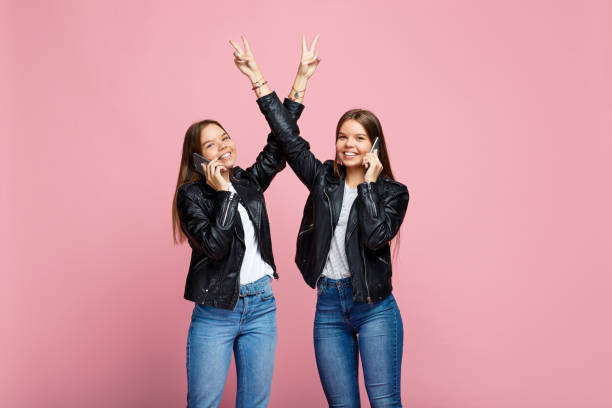 incrível duas jovens gémeas com sorriso brilhante falam em smartphone com levante a mão o gesto de paz mostrar sobre fundo rosa. - multiple birth - fotografias e filmes do acervo