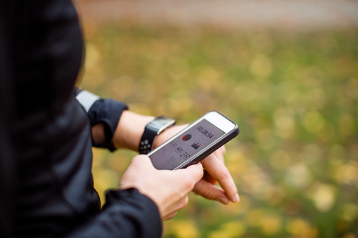Close up of a urban runner using sporty smart watch and smartphone app for training. Jogger syncing her smart watch with smartphone during training outdoors.