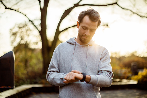 Man wearing sweatshirt standing at the park in morning and using smartwatch. Caucasian fitness male checking heart rate on smart watch app after workout outdoors.