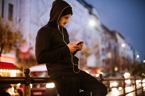 Young man standing outdoors using smart phone and listening to music. Urban runner relaxing after workout in the city. Man wearing black hoodie leaning to a railing by the street.