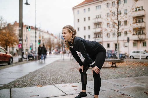 Female runner taking a break during her jog standing on a street. Woman athlete relaxing after workout, resting her hands on her knees.