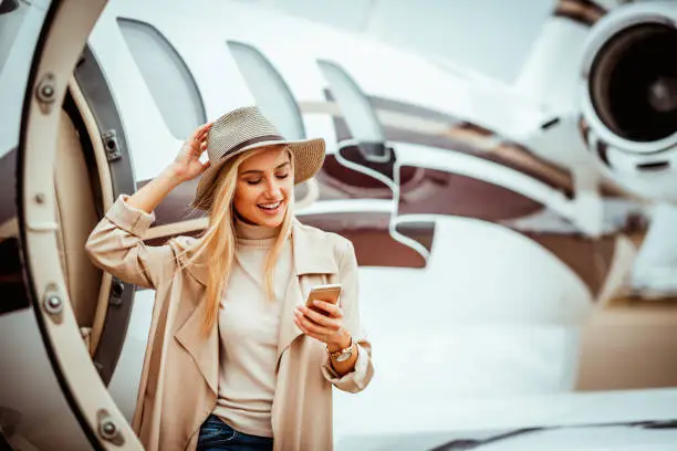 Photo of Rich young woman using her mobile phone while exiting a private jet parked on an airport tarmac