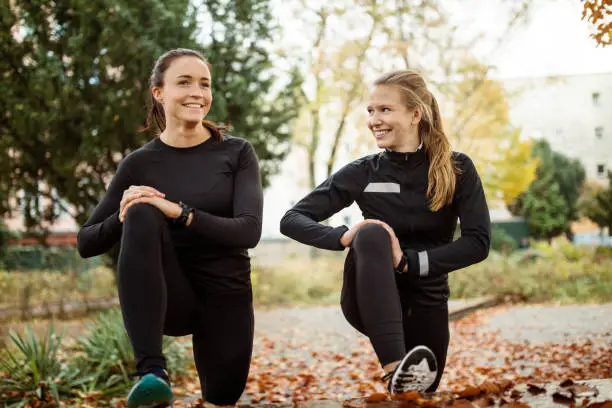 Two young women resting after running workout and stretching legs outdoors. Female runners taking a break after sports training at autumn park.