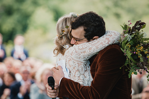 A front view shot of a newlywed couple hugging each other on their wedding day, the bride is wearing a white wedding dress and the groom is wearing a burgundy tuxedo as they celebrate their love for each other.