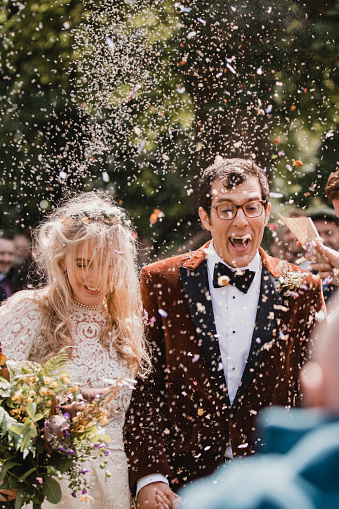 A front view shot of a newlywed couple arm in arm, they are walking down the aisle together on their wedding day, confetti can be seen falling around them as they celebrate their special day.