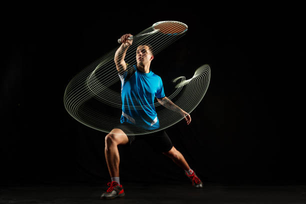 Young male badminton player over balck background Young man playing badminton over black studio background. Fit male athlete isolated on dark with led light trail . badminton player in action, motion, movement. attack and defense concept badminton racquet stock pictures, royalty-free photos & images