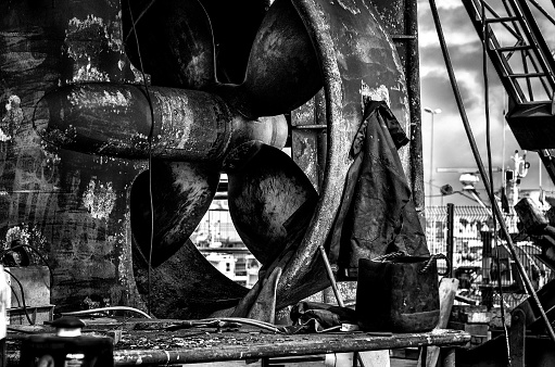 Close-up view of the propeller of a trawler under repair in a shipyard in the town of Guilvinec in Brittany (France) \nHouses can be seen through the propeller\nA worker's coat hangs from the propeller\nThe photo set in black and white