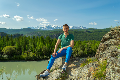 A young man sits on a mountain and smiles on a stone in a green T-shirt, blue pants and sneakers, against the backdrop of a landscape of snow-capped mountains, trees and a river.