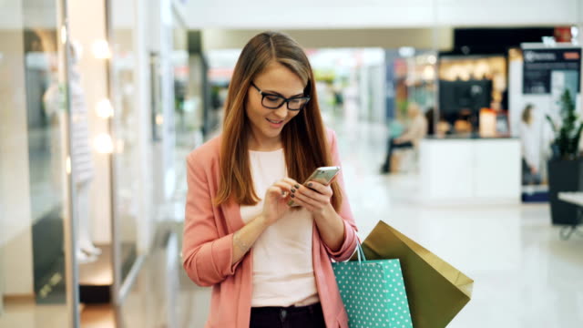 Cherful young woman is using smartphone in shopping center touching screen then looking around at new collection of clothing holding paper bags. Youth and gadgets concept.