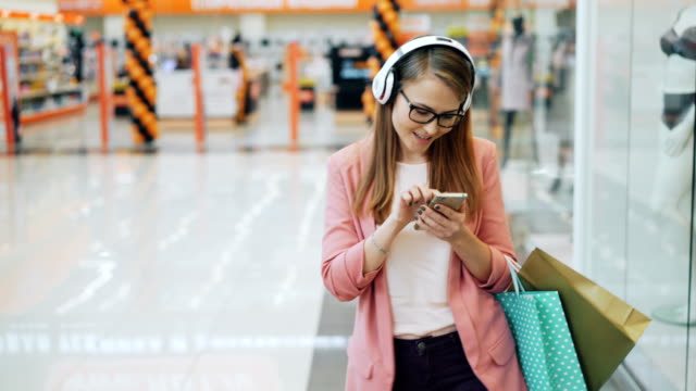 Beautiful lady in headphones is listening to music and using smartphone walking in shopping mall with paper bags. Modern gadgets, millennials and happiness concept.