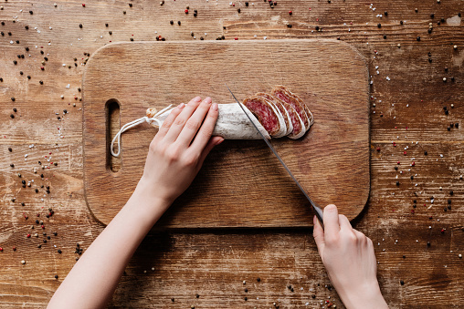 cropped view of woman cutting delicious salami with steel knife on wooden board