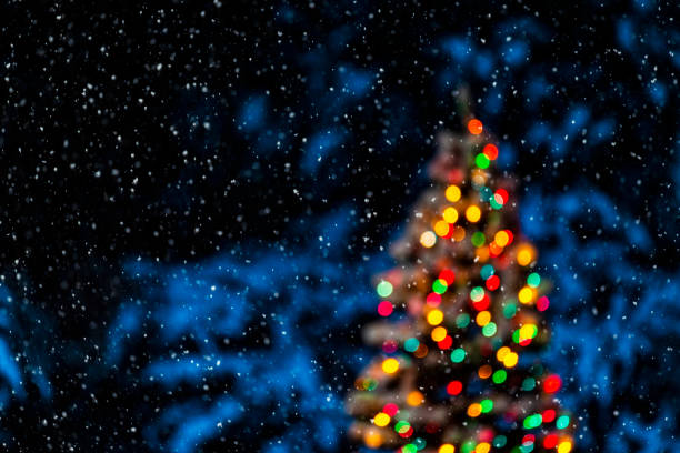 As the snow falls at night with an out of focus Christmas tree with it's lights glowing in the background