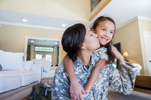 Adult, Hispanic mother in uniform kisses the cheek of her smiling daughter. The little girl has her arms wrapped around her mother's neck in affection, while in their home.