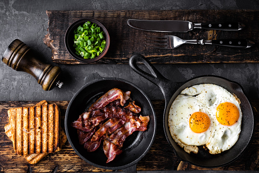 breakfast or brunch fried bacon and eggs in black skillets and crispy toasts top view