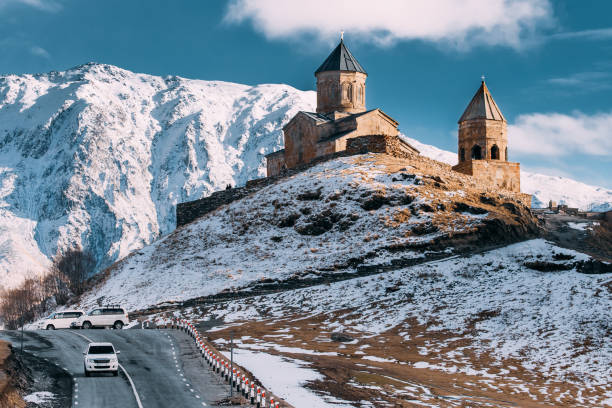 istock Stepantsminda, Georgia. Famous Gergeti Trinity Tsminda Sameba Church In Early Winter Landscape. Suv Moving Near Church In Beautiful Georgian Landscape 1080359260