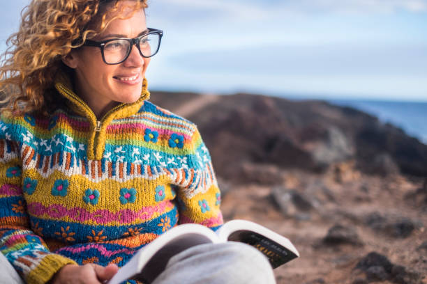 woman sit down on the rocks in outdoor beautiful place with ocean in background reading a paper book and enjoying the freedom and the relax situation. smile and happy people concept - woman with glasses reading a book imagens e fotografias de stock