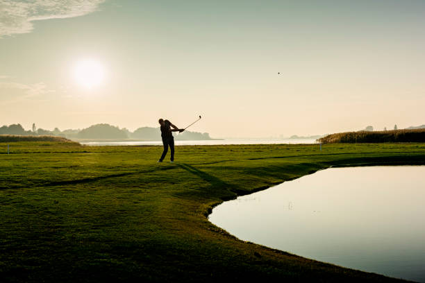 Hitting the perfect pitch shot. Golfer hitting a delicate wedge shot over a water feature and towards the flag. Photographed in the late autumn light on a course on the island of Møn in Denmark. Colour, horizontal with some copy space. taking a shot sport stock pictures, royalty-free photos & images