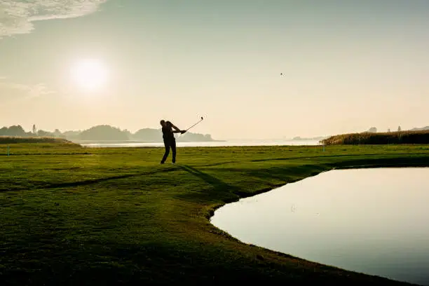 Golfer hitting a delicate wedge shot over a water feature and towards the flag. Photographed in the late autumn light on a course on the island of Møn in Denmark. Colour, horizontal with some copy space.