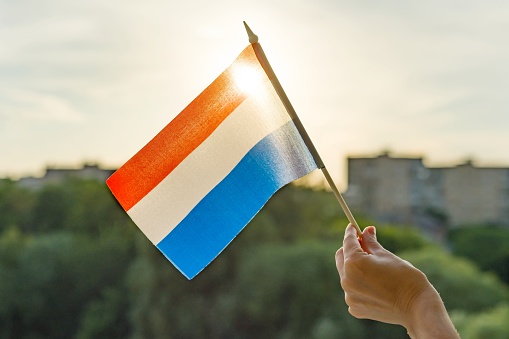 Mano sosteniendo la bandera de Países Bajos una ventana abierta. Cielo azul de fondo, silueta de la ciudad, puesta del sol photo