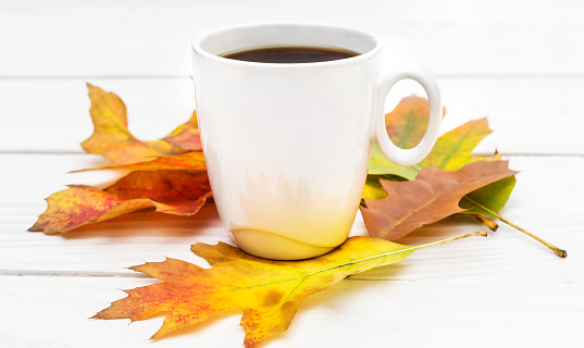Cup of coffee with autumn leaves on white wooden table.