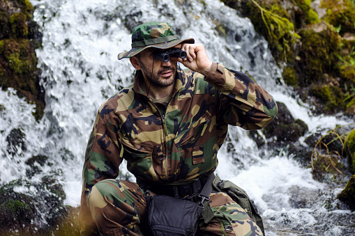 The soldier or park ranger in military uniform is looking away with binoculars on waterfall background.