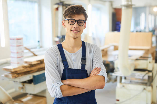 Smiling young joiner in uniform Smiling confident handsome young joiner in uniform standing in modern workshop and crossing arms on chest while looking at camera carpenter portrait stock pictures, royalty-free photos & images