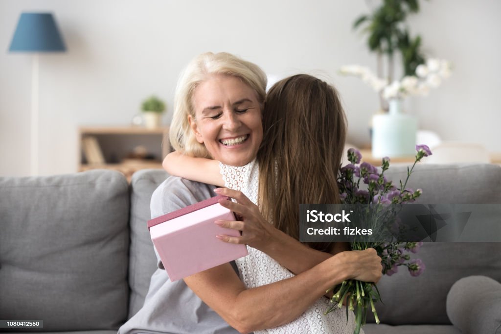 Happy senior grandma hugging granddaughter thanking for gift and flowers Happy senior grandma hugging granddaughter thanking for present holding flower bouquet, smiling excited old grandmother embracing little grandchild girl congratulating granny giving birthday gift box Grandmother Stock Photo