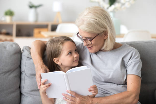 любящая бабушка учит внучку читать книги, сидя на диване - grandparent family reading inside of стоковые фото и изображения