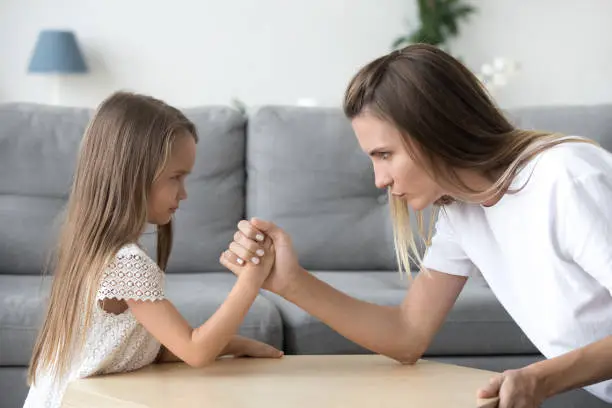 Mom and kid daughter arm wrestling having fight confrontation or family conflict, mother in law and stubborn child girl holding hands competing with serious focused faces expressing disagreement