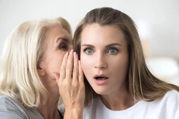 Photo of Older mother whispering in young daughter ear telling shocking news
