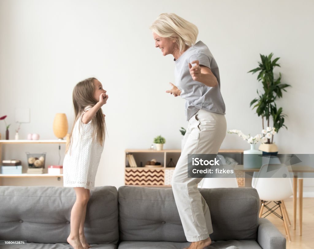 Cute little girl playing with grandmother jumping on couch together Cute little girl having fun playing with smiling grandmother jumping on couch together, happy granny and active kid grandchild dancing on sofa, grandma and granddaughter laughing playing at home Grandmother Stock Photo