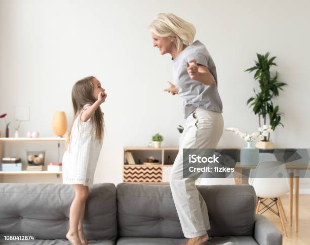 Cute Little Girl Playing With Grandmother Jumping On Couch Together Stock Photo - Download Image Now