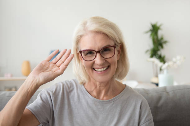 Smiling middle aged woman waving hand looking at camera, portrait Smiling middle aged woman waving hand looking at camera, older mature lady in glasses making video blog or call at home, happy friendly senior vlogger sitting on sofa dating online, headshot portrait waving gesture stock pictures, royalty-free photos & images