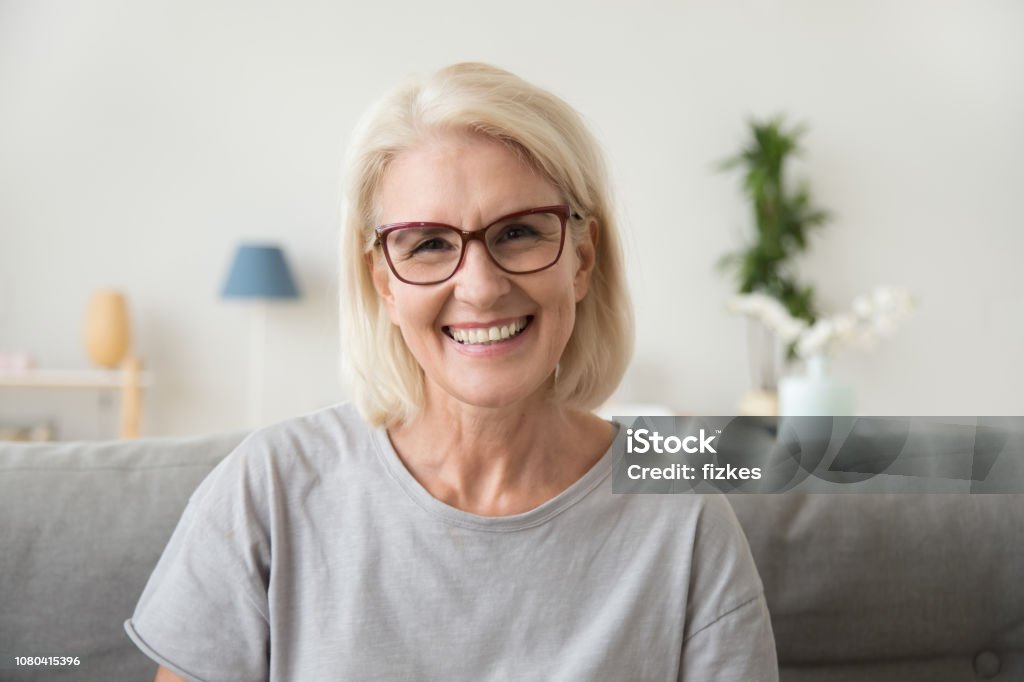 Smiling middle aged mature grey haired woman looking at camera Smiling middle aged mature grey haired woman looking at camera, happy old lady in glasses posing at home indoor, positive single senior retired female sitting on sofa in living room headshot portrait Smiling Stock Photo