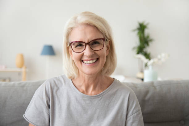Smiling middle aged mature grey haired woman looking at camera Smiling middle aged mature grey haired woman looking at camera, happy old lady in glasses posing at home indoor, positive single senior retired female sitting on sofa in living room headshot portrait midsection photos stock pictures, royalty-free photos & images