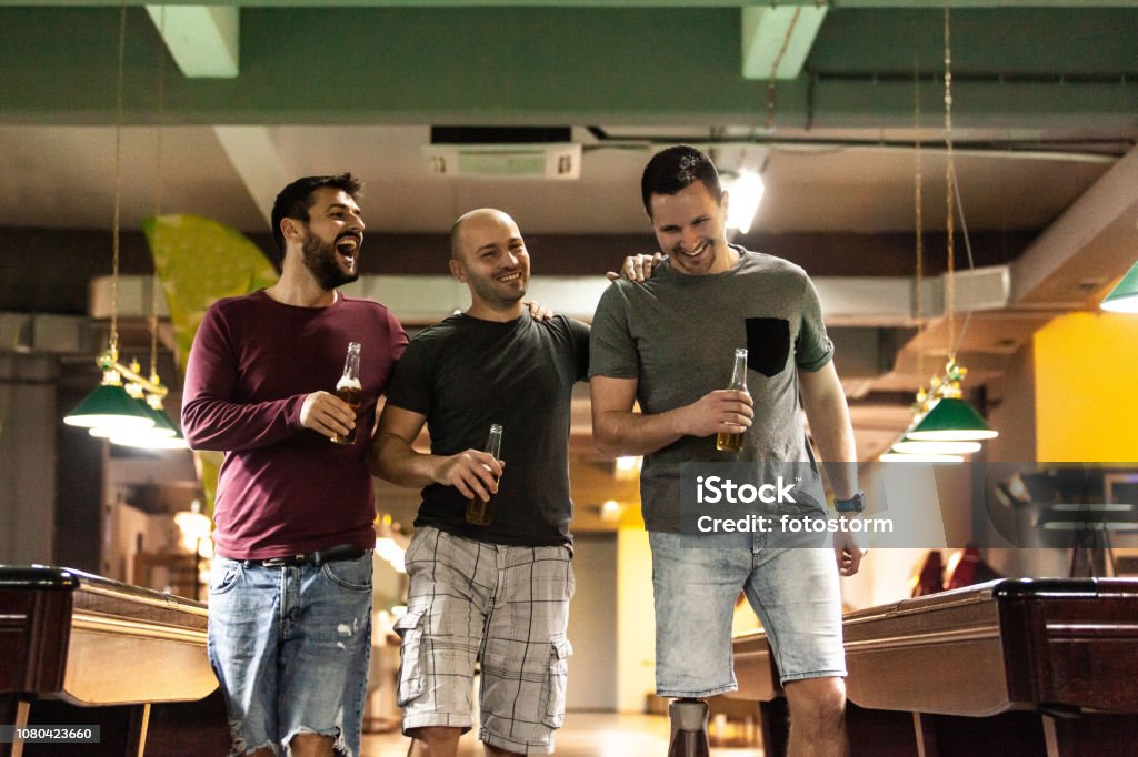 Friends drinking beer in bowling alley Three male friends are having fun in the bowling alley. They are drinking beer and enjoying. Adult Stock Photo