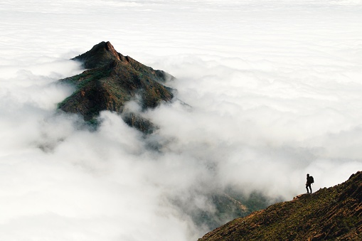 A single person stands on a mountain slope in the distance, looking out over a cloudscape through which a lush mountain peak rises.