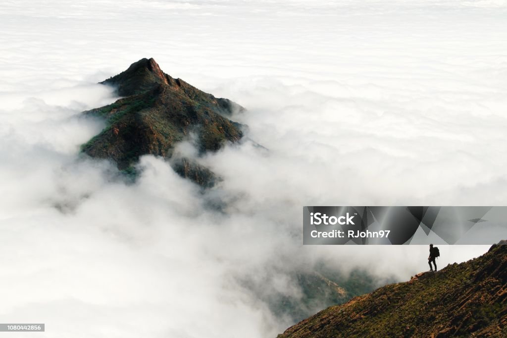 An adventurous male traveler looks out over a mountain peak rising above a thick layer of clouds in Chile's Parque Nacional La Campana A single person stands on a mountain slope in the distance, looking out over a cloudscape through which a lush mountain peak rises. Mountain Stock Photo