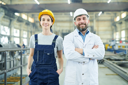 Waist up portrait of two smiling factory workers looking at camera while posing in industrial workshop
