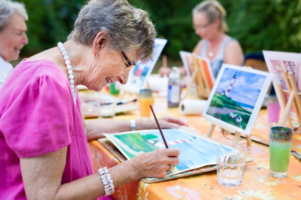 Senior woman smiling while drawing with the group. Side view of a happy senior woman smiling while drawing as a recreational activity or therapy outdoors together with the group of retired women. recreation stock pictures, royalty-free photos & images