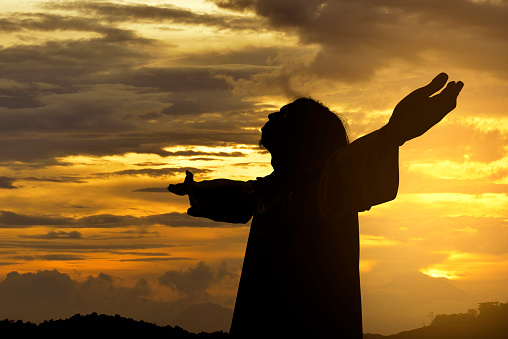 Silhouette of Jesus christ standing with raised arms at sunset background