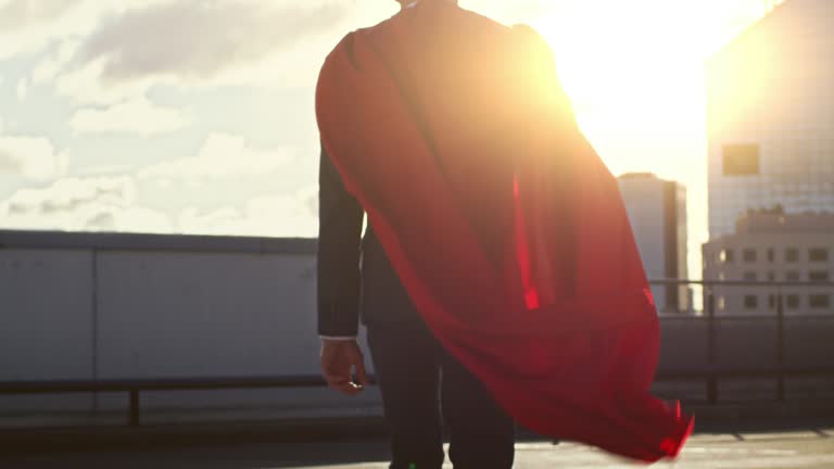 Businessman Superhero WIth Red Cape Blowing in the Wind Walks on the Roof of a Skyscraper, Looking into the Sunset, Ready to Save the Day. Following Back View Slow Motion Shot.