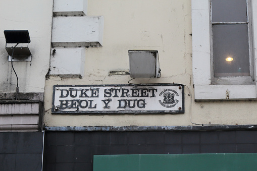 Duke Street name sign written in capital black letters with on a white background on a white and grey wall full of dirt in Cardiff, United Kingdom