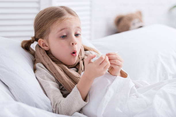 istock shocked sick child with scarf over neck lying in bed and checking temperature with thermometer at home 1080819920