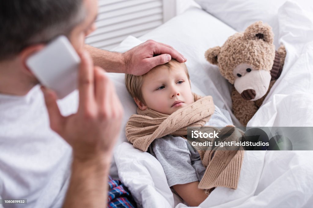 erhöhte Ansicht von Daddy per Smartphone sprechen und berühren kranken Sohn Stirn im Schlafzimmer - Lizenzfrei Krankheit Stock-Foto