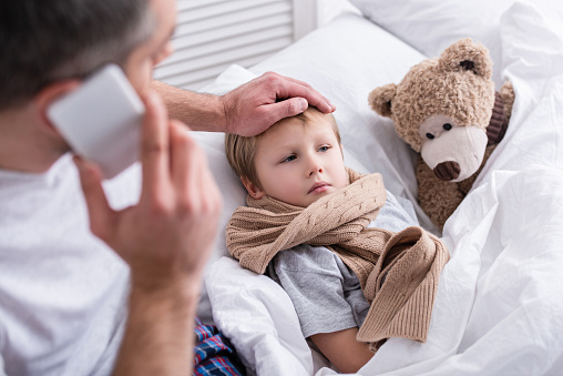 high angle view of daddy talking by smartphone and touching sick son forehead in bedroom