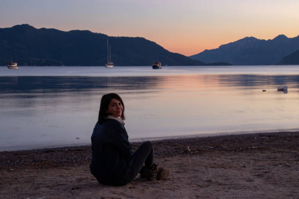 An image of a girl who is sitting on the beach with the sea and mountains on the background at sunset time.