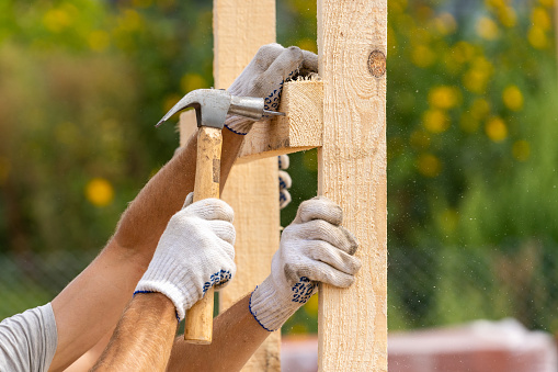 Close up cropped photo of two workers in white protective gloves holding plank board installing it on special house hold construction using hammer and nails
