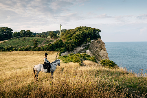 Young woman on horseback admiring the view from the top of the cliffs at Møns Klint in Denmark in the late afternoon light. She is riding a mottled grey horse and she is wearing a long sleeved  t-shirt, riding trousers, riding boots and a protective helmet. Colour, horizontal format with lots of copy space. The tower in the background was part of a NATO early warning radar network that could look across the Baltic towards Russia. Now we are told it monitors shipping passing through the Baltic.