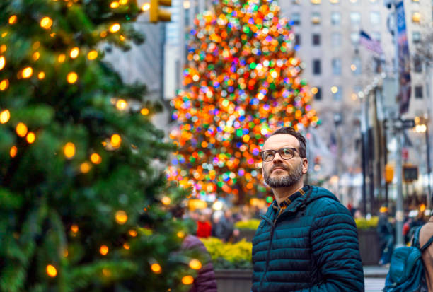 Mature man looking away while standing in city. Male is celebrating Christmas. He is in warm clothing against illuminated trees.